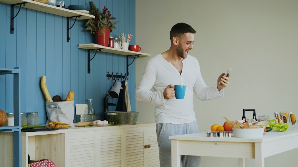 An adult man glancing at his phone while holding a mug beside the kitchen table at home