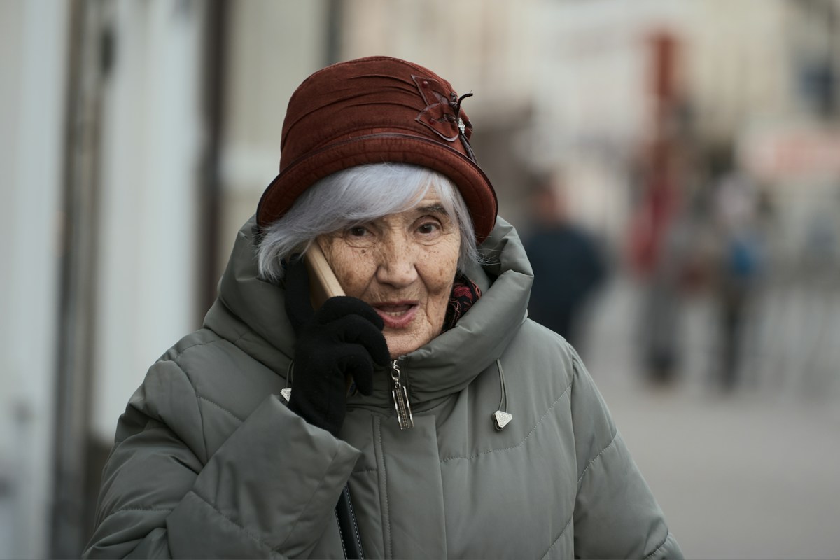 An older adult woman in a gray coat enjoying a phone conversation while outdoors on a quiet street