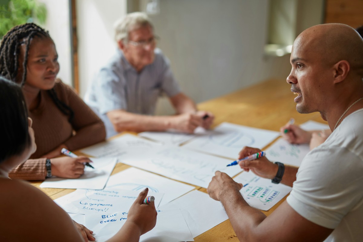 A diverse group of adult colleagues, including an older man, collaborating on handwritten notes around a sunlit table
